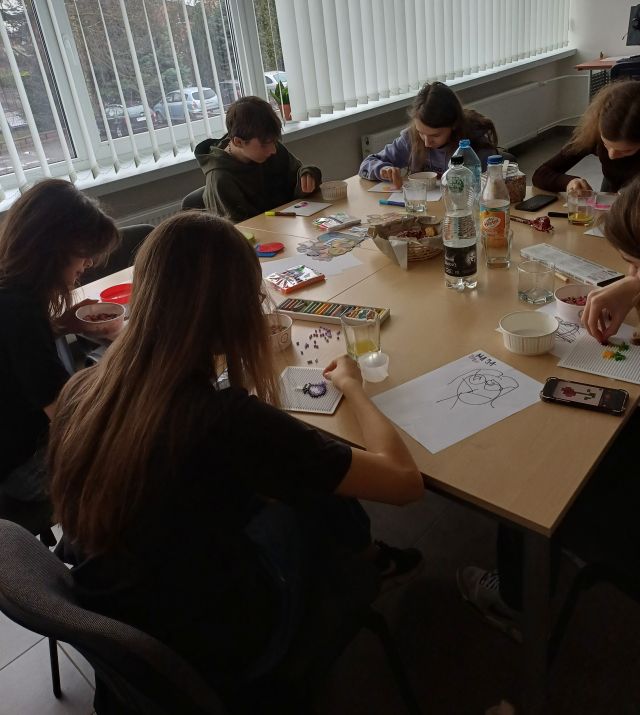 młodzież siedzi przy stolikach i układa koraliki