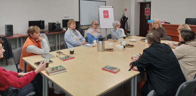 9 kobiet siedzi przy połączonych stołach, w tle komputery