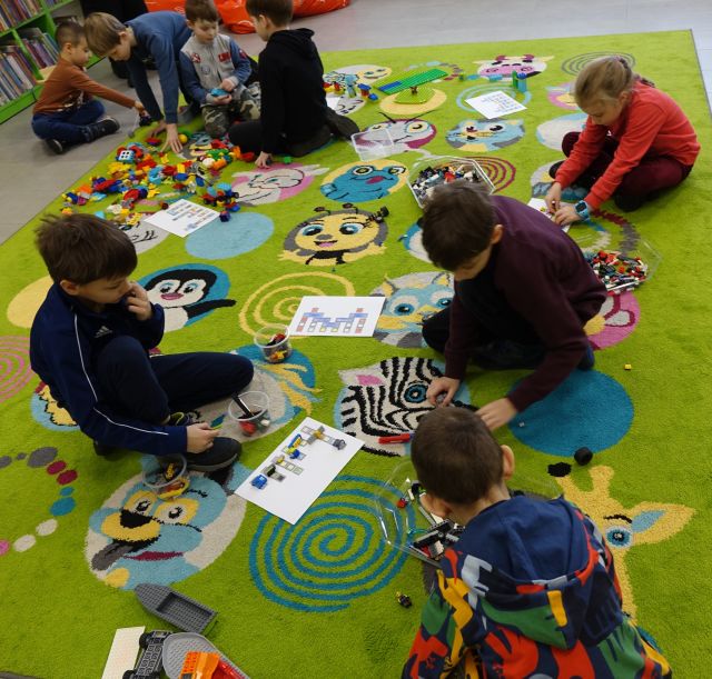 dzieci siedząc na kolorowym dywanie układają klocki LEGO