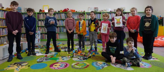 dzieci uczestniczące w warsztatach z LEGO pozują do zdjęcia, w tle regały z książkami
