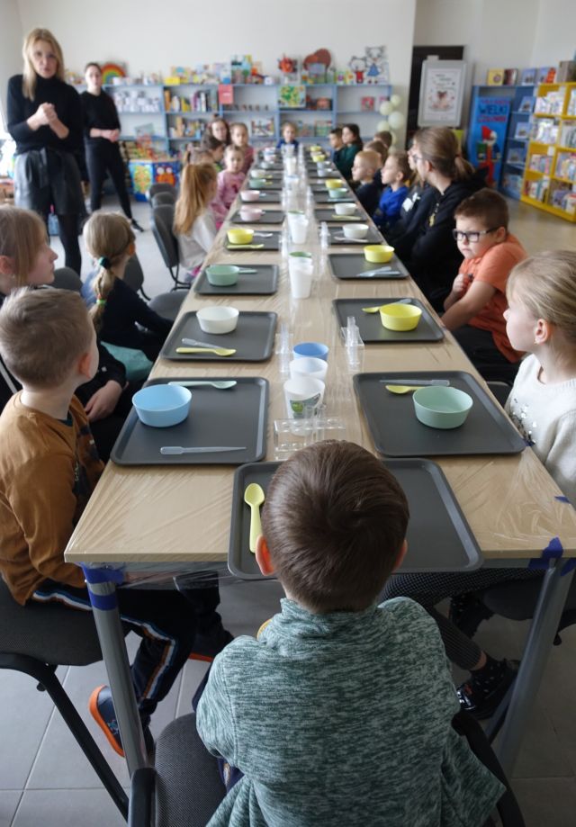 25 dzieci siedzi przy stołach, na których na tackach stoją plastikowe miseczki i łyżeczki, panie które stoją opowiadają, w tle regały z książkami