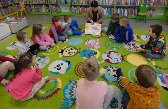 przedszkolaki i bibliotekarka siedzą na kolorowym dywanie, w tle regały z  książkami