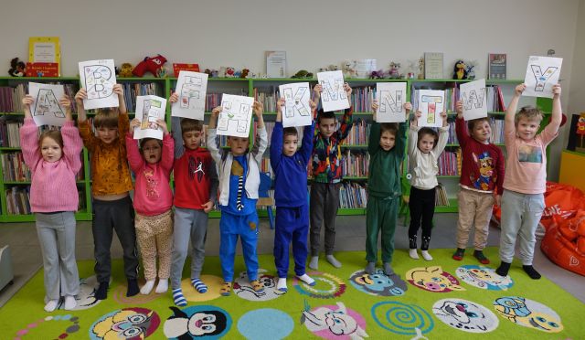 przedszkolaki pozują do zdjęcia trzymając wysogo nad głowami w rękach pokolorowane przez siebie literki, w tle regały z książkami