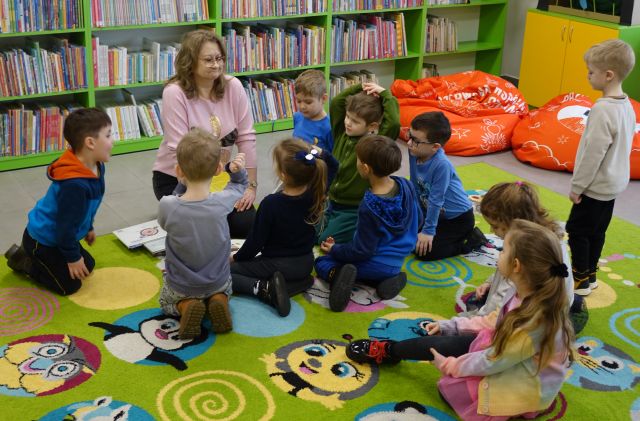 przedszkolaki siedzą na kolorowym dywanie z bibliotekarką, w tle regały z książkami