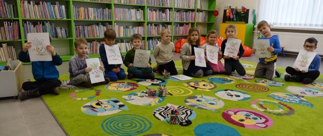 przedszkolaki siedzą na kolorowym dywanie i pozyją do zdjęcia z wykonanymi przez siebie rysunkami na kartkach, w tle zielone regały z książkami
