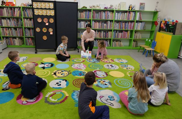 dzieci siedzą na kolorowym dywanie, przed nimi na krzesełku siedzi bibliotekarka, w tle regały z książką