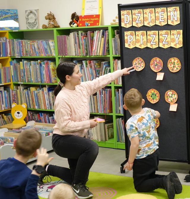 bibliotekarka pokazuje dzieciom pizze namalowane na kartkach i przypięte do czarnej tablicy