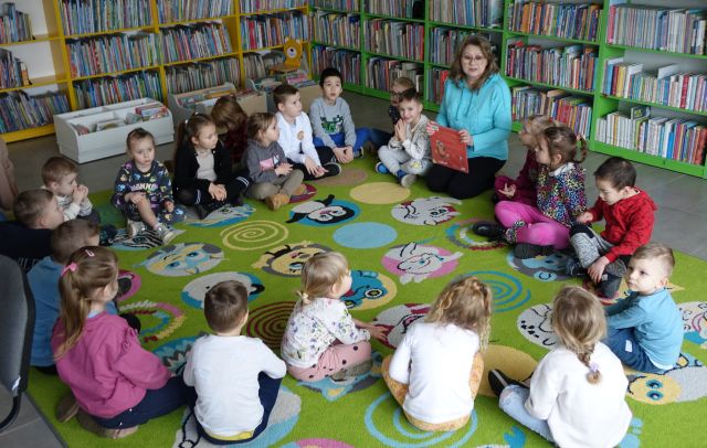 przedszkolaki i bibliotekarka z książką siedzą na kolorowym dywanie, w tle regały z książkami