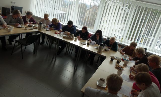 Prawei 20 seniorek siedzi przy stołach ustawionych w podkowę i rozwiązuje zadania