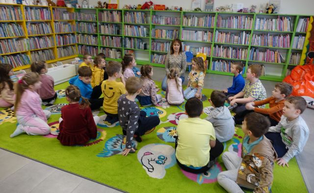 przedszkolaki siedzą na kolorowym dywanie, przed nimi siedzi bibliotekarka, w tle regały z książkami