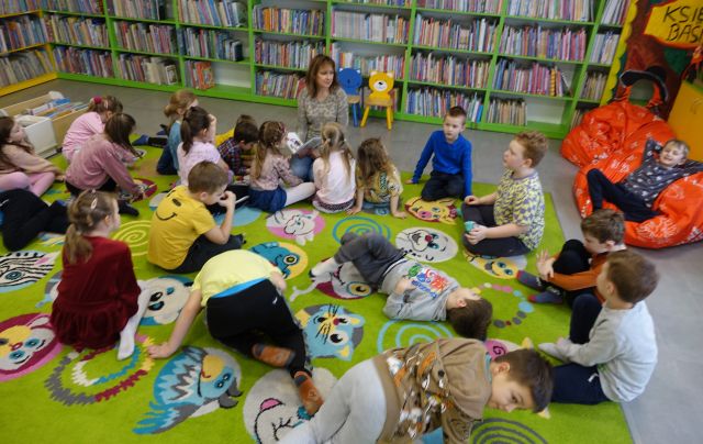 przedszkolaki siedzą na kolorowym dywanie, przed nimi bibliotekarka z książką, w tle regały z książkami