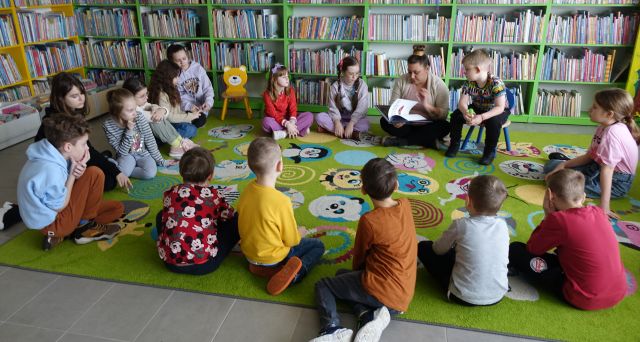 bibliotekarka czyta dzieciom siedzącym na kolorowym dywanie książkę, w tle regały z książkami