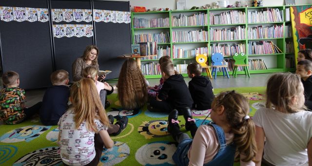 dzieci siedzą na kolorowym dywanie i słuchają czytającej bibliotekarki, w tle regały z książkami