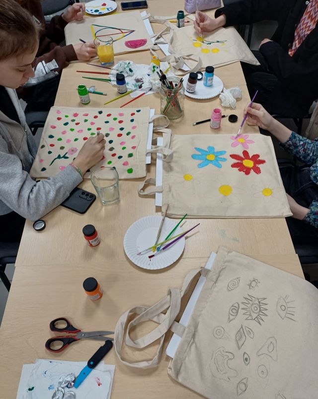 młodzież siedząca przy stołach maluje torby