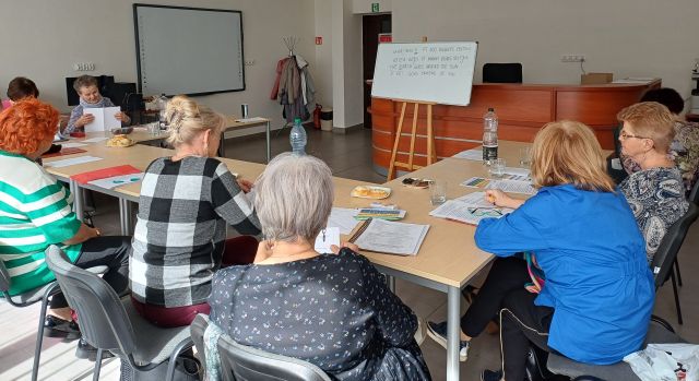 seniorki siedzą przy stołach, na stołach leżą kartki i zeszyty, w tle tablica