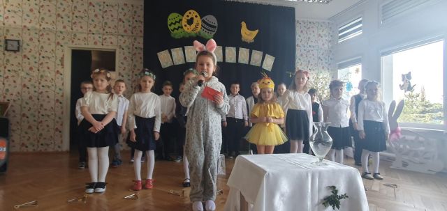 dziewczynka w stroju zająca trzyma mikrofon i kopertę, za nią stoją przedszkolaki