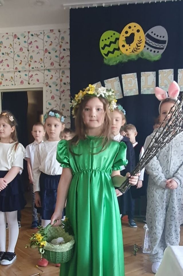 dziewczynka w zielonej sukience z baziami i koszyczkiem, za nią stoją przedszkolaki
