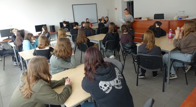 bibliotekarka stoi z kartka przed siedzącą przy stolikach młodzieżą, w tle komputery i tablica interaktywna