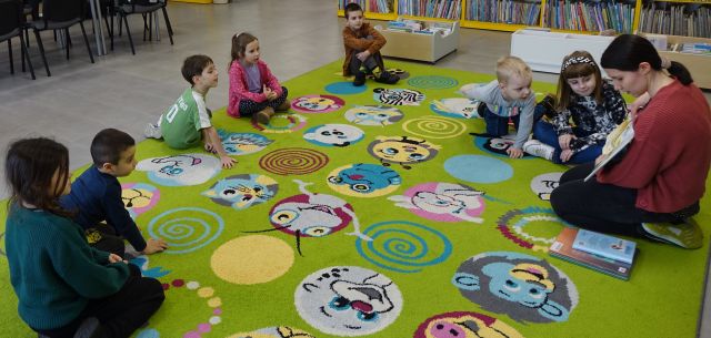 dzieci i bibliotekarka z książką siedzą na kolorowym dywanie, w tle regały z książką
