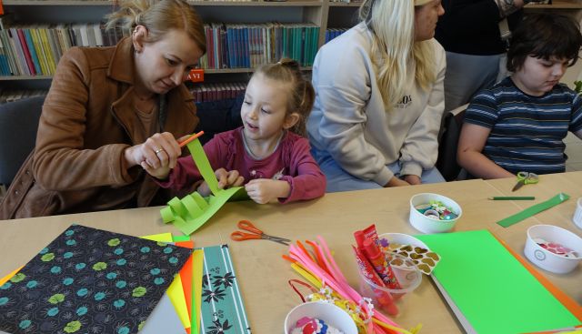 dzieci i rodzice wykonują kwiatki z zielonych kartek, w tle regały z książkami