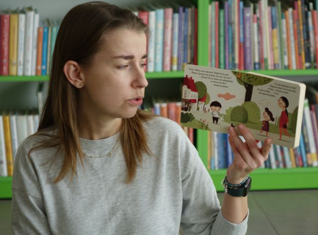 kobieta pokazuje dzieciom książkę, w tle regały z książkami