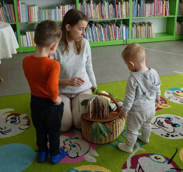 kobieta pokazuje małym dzieciom koszyk z warzywami, w tle regały z książkami