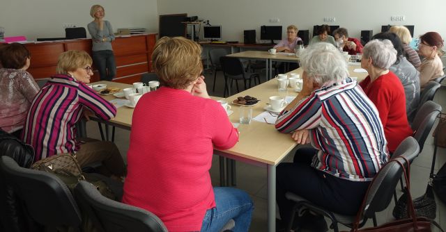 seniorki siedzące przy stołach ustawionych w podkowę słuchają stojącej przed nimi opartej o biurko kobiety
