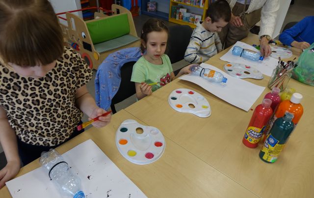 dzieci siedzące przy stołach malują różnymi kolorami plastikowe butelki