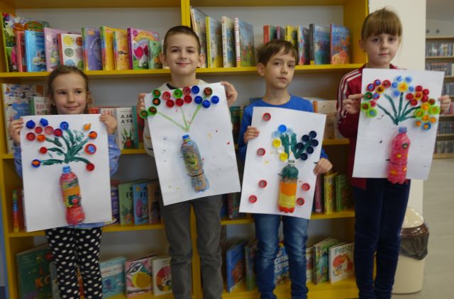 4 dzieci prezentuje swoje prace plastyczne przedstawiające kwiaty z pomalowanych plastikowych butelek i nakrętek, w tle regały z książkami