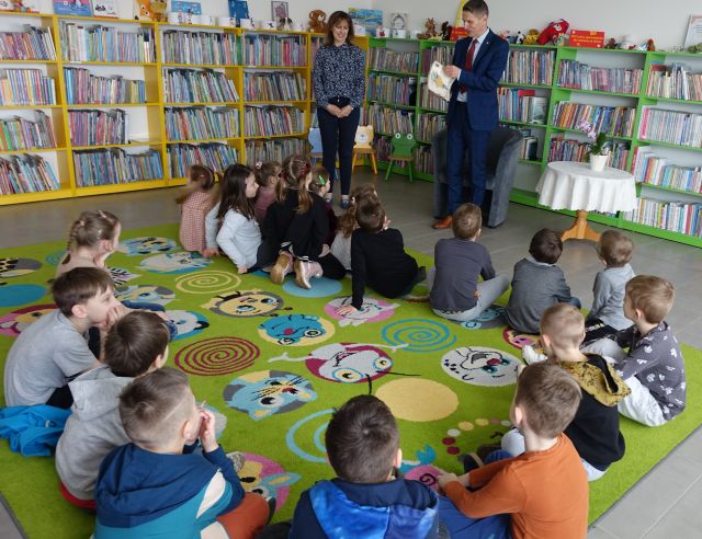 dzieci siedzą na kolorowym dywanie, przed nimi stoi dyrektorka biblioteki i burmistrz, w tle regały z książkami