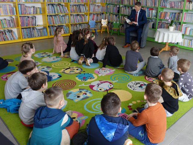 dzieci siedzą na kolorowym dywanie, przed nimi na siwym fotelu siedzi burmistrz, w tle regały z ksiązkami