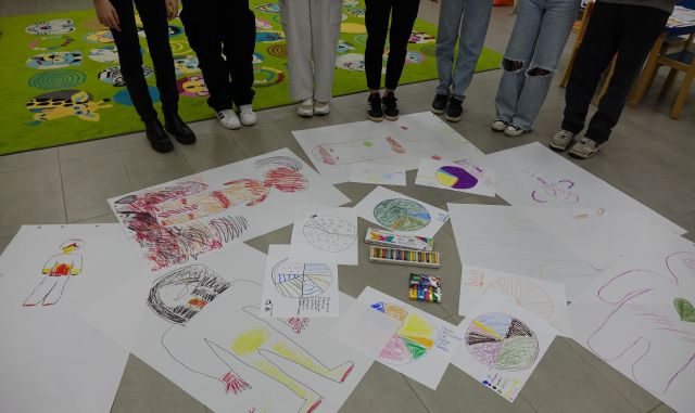 na podłodze rozłożone rysunki młodzieży, przed rysunkami widać stopy 7 osób