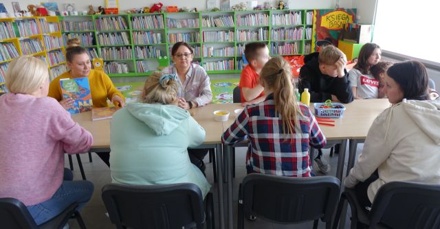młodzież siedzi przy połączonych stołach, bibliotekarka pokazuje jej książki, w tle regały z książkami