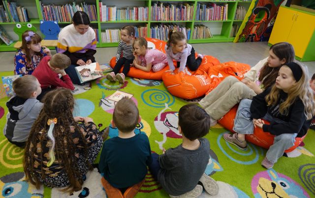 dzieci siedzą na kolorowym dywanie i pomarańczowych pufach, przed nimi siedzi bibliotekarka z książką, w tle regały z książkami