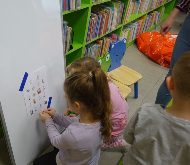 dwie dziewczynki przyczepiają do tablicy magnetycznej obrazki, w tle regały z książkami