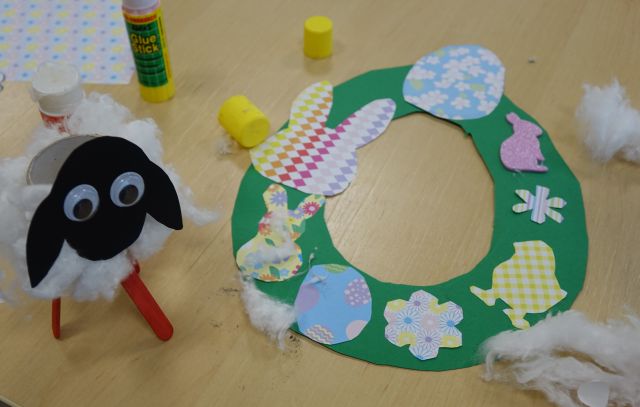 na zielonej obręczy naklejone wycięte z kartek króliki, jajka i kwiaty, obok baranek zrobiony z rolki po papierze toaletowym i waty