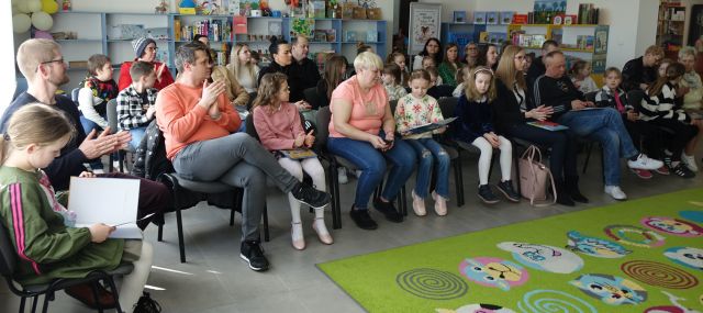 27 dzieci z rodzicami i nauczycielami siedzi na krzesłach, w tle kolorowe regały