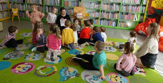 bibliotekarka pokazuje siedzącym dzieciom książki, w tle regały z książkami