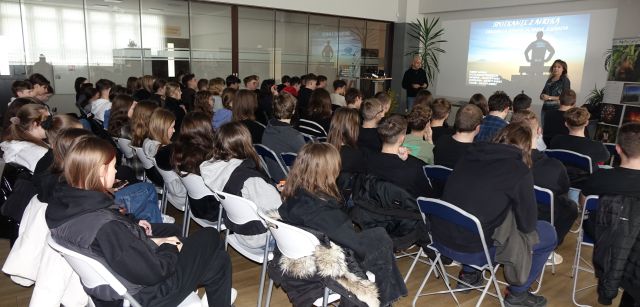 młodzież uczestnicząca w spotkaniu z podróżnikiem siedzi na krzesłach, przed nimi gość i dyrektorka biblioteki, w tle wyświetlana prezentacja
