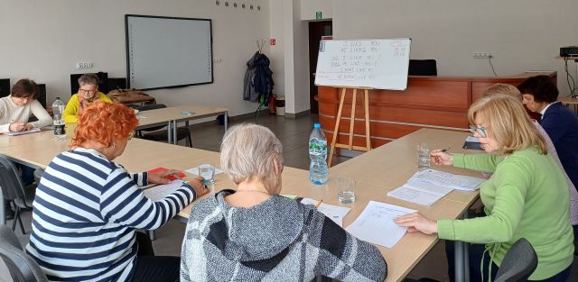 seniorki siedzą przy stolikach i notują na kartkach, przed nimi tablica i biurko, w tle komputery