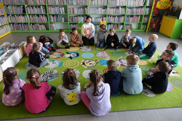 bibliotekarka z książką siedzi na kolorowym dywanie, obok w okręgu siedzą przedszkolaki, w tle kolorowe regały z książkami