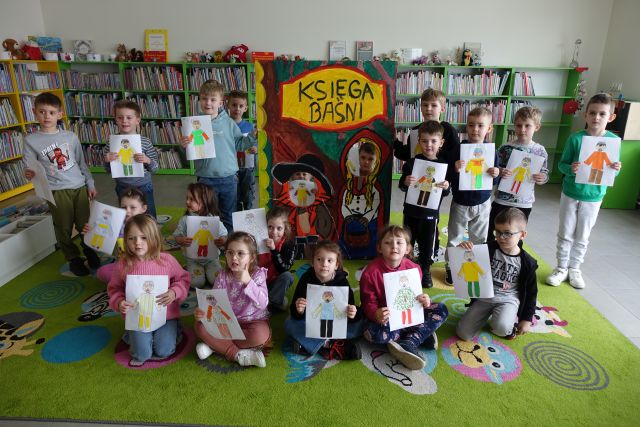 przedszkolaki pozują do zdjęcia z wykonanymi przez siebie wyklejonymi postaciami cesarzy, w tle fotobudka Księga baśni i kolorowe regały z książkami