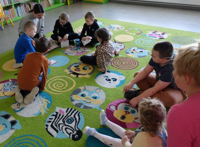 dzieci siedzące na dywanie patrzą na bibliotekarkę trzymającą książkę, w tle regały z książkami
