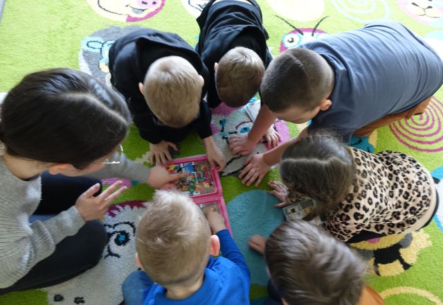 dzieci i bibliotekarka siedzące na kolorowym dywanie pochylają się nad książką