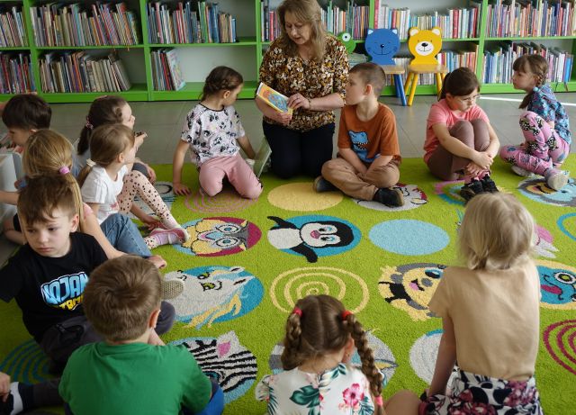 dzieci i bibliotekarka z książką siedzą w okręgu na kolorowym dywanie, w tle regały z książkami