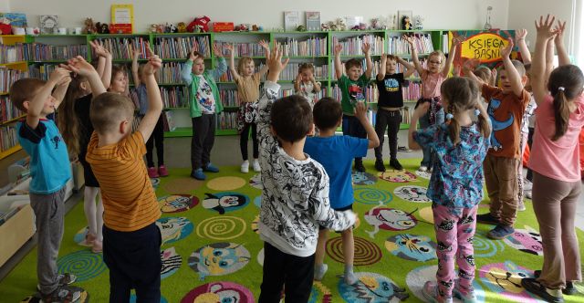 dzieci w kółku stoją z podniesionymi do góry rękoma, w tle regały z książkami