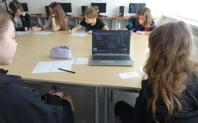 młodzież siedzi przy stołach i rozwiązuje zadania matematyczne, na stole stoi włączony laptop
