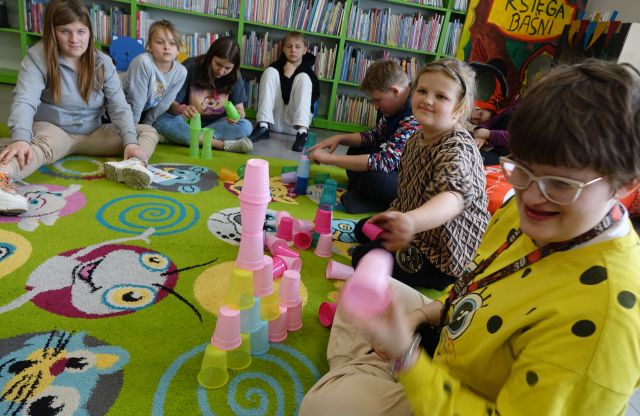 młodzież siedząca na kolorowym dywanie układa kolorowe kubeczki w wieżę, w tle regały z książkami