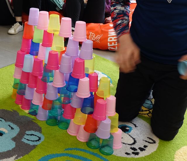 kolorowe kubeczki ułożone jeden na drugim, z prawej strony dłonioe i nogi mężczyzny