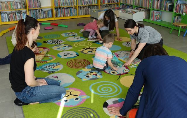 dzieci bibliotekarka i mamusie i jeden tatuś siedzą na kolorowym dywanie i przeglądają książki